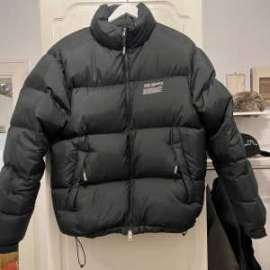 Svart pufferjacka från Axel Arigato - Säljer en snygg svart pufferjacka från Axel Arigato. Jackan har en dragkedja framtill och två fickor med dragkedjor. Perfekt för kalla dagar med sin tjocka vaddering. Den har en stilren design med märkets logga på bröstet. Passar både till vardags och mer uppklädda tillfällen.
