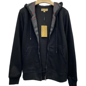 Svart hoodie från Burberry - Säljer en snygg svart hoodie från Burberry med dragkedja och klassiskt rutigt mönster i luvan. Den har långa ärmar och en broderad logga på bröstet. Perfekt för höst och vår! Alla storlekar finns