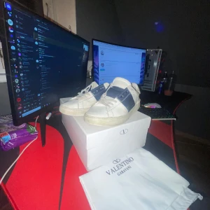 Valentino Garavani Open - Säljer ett par snygga Valentino Garavani sneakers i vitt och blått. De är i bra skick och kommer med originalförpackning och dustbag. Perfekta för både vardag och fest! Skorna har en stilren design med en bred blå rem över ovansidan.kommer såklart fixa till de och städa de.