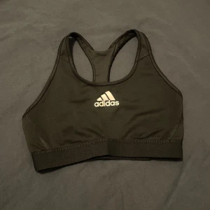 Svart sporttopp från Adidas - Säljer en svart sporttopp från Adidas i mycket bra skick. Den är perfekt för träning med sin bekväma passform och stödjande design. Toppen har en klassisk brottarrygg och det ikoniska Adidas-loggan på framsidan. Perfekt för gymmet eller löprundan!