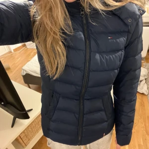 Mörkblå dunjacka från Tommy Hilfiger - Säljer en snygg mörkblå dunjacka från Tommy Hilfiger. Jackan har en mysig pälskantad huva och dragkedja framtill. Perfekt för kalla vinterdagar! Den är i mycket bra skick och passar både till vardags och lite finare tillfällen❤️är i storlek M men passar mer som en S.