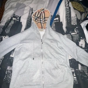 Burrberry hoodie - Säljer en snygg grå hoodie med dragkedja och en klassisk rutig mönstrad insida i huvan. Perfekt för höst och vår, med långa ärmar och en bekväm passform. Den har praktiska fickor framtill och är i bra skick. Perfekt för en casual look!