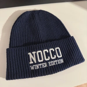 Nocco mössa - Säljer en snygg mörkblå  mössa från noccos winter edition. Perfekt för kalla dagar med stilren design och broderad text framtill. Håller dig varm och ser cool ut samtidigt!