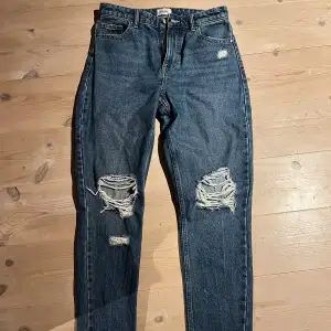 Säljer ett par snygga blå jeans med coola slitningar på knäna. De har en klassisk femficksdesign och en vanlig gylf. Perfekta för en avslappnad och trendig look. Passar bra till både vardag och fest!