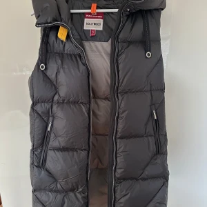 Svart dunväst från Parajumpers - Säljer en svart dunväst från Parajumpers i mycket bra skick. Västen har en dragkedja framtill och praktiska fickor med dragkedjor. Perfekt för höst och vinter, håller dig varm och stilren. Passar både till vardags och mer avslappnade tillfällen. Den är helt oanvänd med alla prislappar på. 