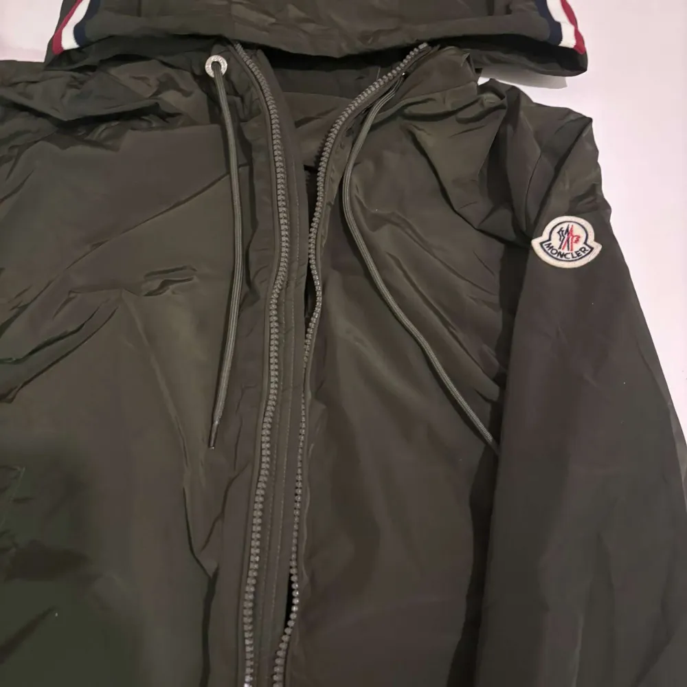 Säljer en snygg grön vindjacka från Moncler. Jackan har en dragkedja framtill och en huva med röd och vit randig detalj. Perfekt för höst och vår! Den är i mycket bra skick och passar både till vardags och lite finare tillfällen.. Jackor.