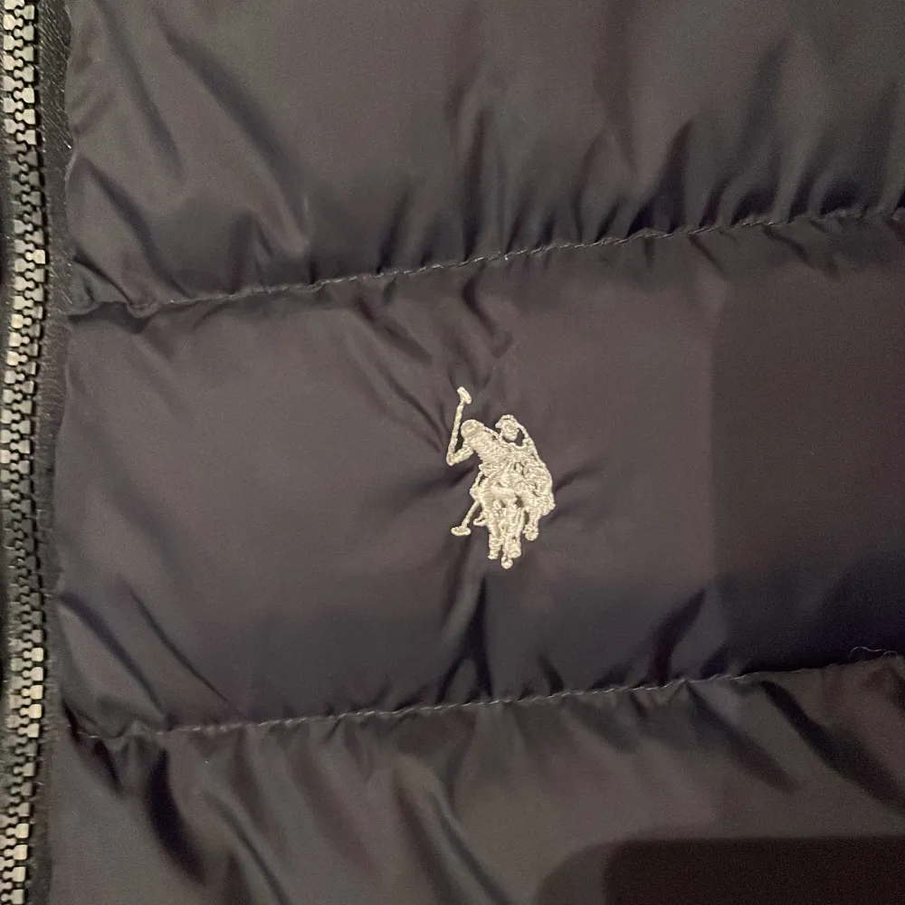 Säljer en snygg svart dunväst från U.S. Polo Assn. i storlek S. Västen har en dragkedja framtill och är quiltad för extra värme. Perfekt för höst och vår! Den har en broderad logga på bröstet och två praktiska fickor med dragkedja.. Jackor.