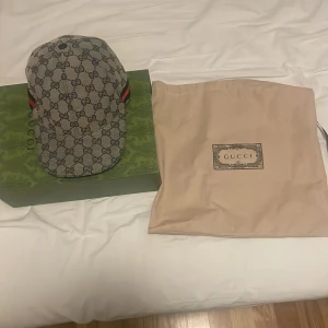 Gucci keps med monogram - Säljer en snygg Gucci keps i storlek M, 57 cm. Den har det klassiska Gucci-monogrammet i grått och svart med röda och gröna detaljer på sidorna. Perfekt för att ge din outfit en lyxig touch. Kepsen kommer med originalförpackning och dustbag.ÄKTA