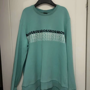 Grön sweatshirt från Boss - Säljer en snygg grön sweatshirt från Boss i storlek M. Tröjan har ett coolt tryck med 'BOSS' i svart och vitt över bröstet. Den är långärmad och perfekt för både höst och vår. Materialet känns mjukt och bekvämt, perfekt för en avslappnad stil. Passar bra till jeans eller joggers!