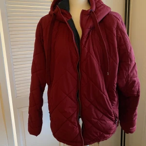 Vinröd quiltad jacka med huva strl 42/44 - Säljer en snygg vinröd quiltad jacka med huva. Jackan har dragkedja framtill och praktiska fickor med dragkedjor. Perfekt för höst och vinter, håller dig varm och stilren. Passar både till vardags och lite finare tillfällen. 🧥