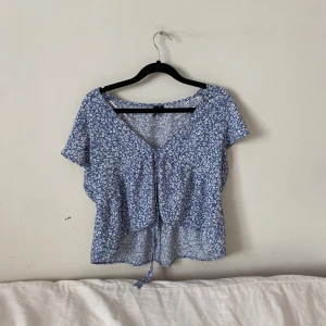 Blå blommig blus - Söt blå blus med vita blommor, perfekt för sommarn. Den har korta ärmar och en knytning framtill som ger en fin detalj. Blusen är i ett lätt och luftigt material, vilket gör den idealisk för varma väder. Passar både till vardags och festligare tillfällen.