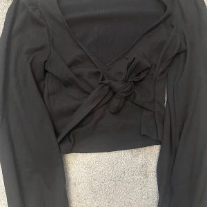 Svart omlottopp från Monki - Säljer en snygg svart omlottopp från Monki i storlek M. Toppen har långa ärmar och en knytning framtill som ger en fin passform. Perfekt för både vardag och fest!