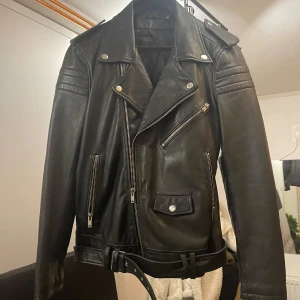 Svart skinnjacka med bälte - Säljer en snygg svart skinnjacka i klassisk bikerstil. Jackan har dragkedja, knappar och ett bälte i midjan för extra stilpoäng. Perfekt för höst och vår, och funkar lika bra till fest som till vardags. Den har långa ärmar och flera praktiska fickor med dragkedjor.