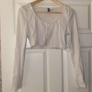Vit croppad topp från H&M Divided - Säljer en vit croppad topp från H&M Divided i storlek M. Toppen har långa ärmar och knappar framtill, perfekt för en stilren look. Den är i bra skick och passar utmärkt till både vardag och fest.