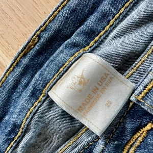 True Religion stl 25 - Säljer ett par snygga blå jeans från True Religion i storlek 25. De har en klassisk femficksdesign med broderade detaljer på bakfickorna och en bekväm passform. Perfekta för både vardag och fest! 🕺OBS! fanns ett hål i grenen som har lagats hos skräddare och syns knappt.
