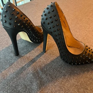 Svarta pumps med nitar - Snygga svarta pumps från Nelly med coola nitar över hela ytan. De har en hög klack och spetsig tå, vilket ger en edgy look. Perfekta för en utekväll eller fest! Skorna är i bra skick och redo att användas för att ge din outfit det lilla extra.
