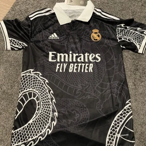 Svart Real Madrid fotbollströja från Adidas - Säljer en helt ny svart Real Madrid fotbollströja från Adidas i nytt skick. Tröjan har ett coolt drakmönster i grått och vitt, med klubbens emblem och sponsorlogga i guld. Den är kortärmad med en vit krage och vita detaljer på ärmarna. Perfekt för alla fans! 🐉⚽