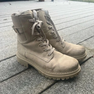 Beige kängor med snörning - Säljer ett par beige kängor i bra skick. De har snörning och dragkedja på sidan för enkel på- och avtagning. Insidan är fodrad med mjukt material för extra värme, perfekt för höst och vinter. Sulan är robust och ger bra grepp. Passar både till vardags och lite tuffare väder. Kängorna är ifrån Rieker och är i storlek 40, går såklart att göra skorna rena🤍