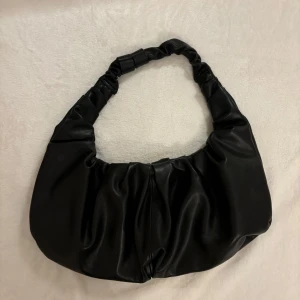 Svart axelväska i syntetmaterial - Snygg svart axelväska i syntetmaterial med en unik, rynkad axelrem. Perfekt för både vardag och fest. Väskan har en stilren design som passar till alla outfits. Den är rymlig nog för att få plats med alla dina nödvändigheter .  Säljer för att den inte kommer till användning 