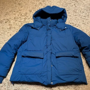 Blå dunjacka från Kenzo - Säljer en snygg blå dunjacka från Kenzo. Jackan har en huva och långa ärmar, perfekt för kalla dagar. Den är i mycket bra skick och har praktiska fickor framtill. Stängs med dragkedja och knappar för extra värme. Perfekt för vintern!