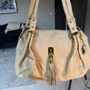Beige handväska - Säljer en snygg beige axelväska i skinn med gulddetaljer och en  frans framtill, som går att ta bort. Bra skick