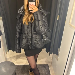 Svart pufferjacka med huva - Säljer en snygg och varm svart pufferjacka med huva. Jackan har en glansig yta och är perfekt för kalla dagar. Den har långa ärmar och en dragkedja framtill. Superbekväm och stilren för vintern!