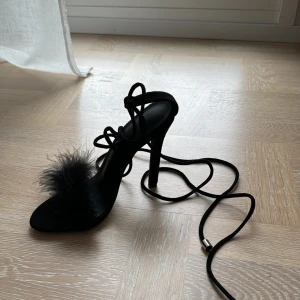 Svarta klackskor med fjäderdetalj - Säljer ett par eleganta svarta klackskor med en unik fjäderdetalj framtill. Skorna har en hög klack och smala remmar som knyts runt vristen för en chic look. Perfekta för en fest eller en utekväll! Helt oanvända! 