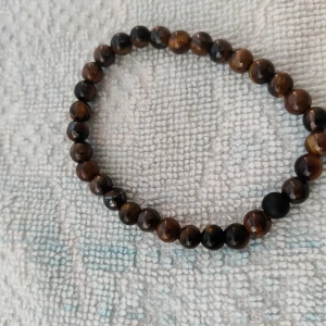 Två elastiska armband med pärlor - Säljer två snygga elastiska armband. Det ena armbandet har bruna och svarta pärlor med en cool tigeröga-effekt, medan det andra är helt svart med en enda brun pärla som detalj. Perfekta för att ge en stilren touch till din outfit!