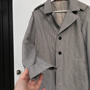 Rutig kappa från Premium - Säljer min pappas stilrena rutiga kappa från Premium. Den har en klassisk design med knappar framtill och en snygg krage. Perfekt för höst och vår. 