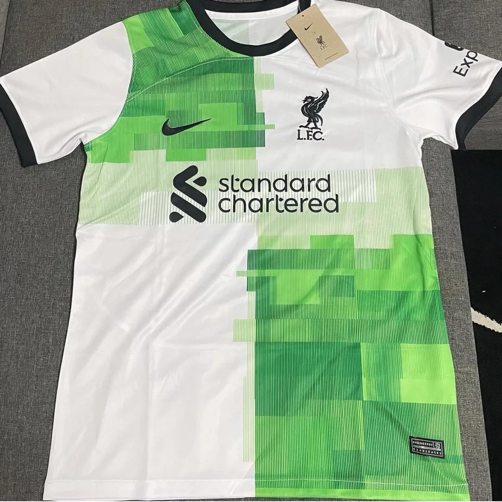 Säljer en snygg vit och grön fotbollströja från Nike med L.F.C.-logga och Standard Chartered som sponsor. Tröjan har korta ärmar och en modern design med pixelmönster i grönt. Perfekt för alla fotbollsfans! finns även i m. T-shirts.