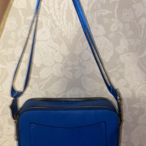 Blå axelväska med dragkedja - Säljer en snygg blå axelväska i syntetmaterial. Väskan har en praktisk dragkedja och en justerbar axelrem. Perfekt för både vardag och fest!