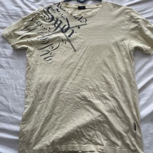 Beige t-shirt med tryck - Säljer en beige t-shirt med ett coolt tryck över bröstet. Den är kortärmad och perfekt för en avslappnad stil. Materialet känns mjukt och bekvämt, perfekt för vardagsbruk. strl XXL men är mindre i storlek
