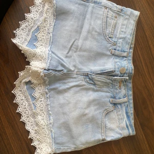 Ljusa jeansshorts med spetsdetaljer - Säljer ett par supergulliga ljusa jeansshorts med vit spetskant längst ner. De har en klassisk femficksdesign och stängs med dragkedja och knapp. Perfekta för sommardagar eller festivaler! 🕶️