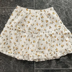 Blommig kortkjol med volang - Säljer en superfin kortkjol med blommigt mönster i gult och grönt på en vit bakgrund. Kjolen har en söt volangdetalj och är perfekt för sommardagar. Den är gjord i ett lätt och bekvämt material, vilket gör den idealisk för både vardag och fest. Passar perfekt med en enkel topp eller en mysig tröja. Storlek XS men passar även S.