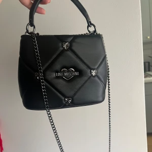 Love Moschino väska - Söt väska från Love Moschino som är köpt på Zalando. Det är en bucket modell som kallas för Jewel heart bag. Man kan ha den crossbody med kedjan eller ta av den och bara ha den som en handväska. Inget slitage alls. Mycket bra skick.