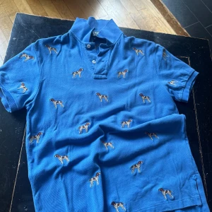 Blå pikétröja med hundmotiv från Polo Ralph Lauren - Säljer en snygg blå pikétröja från Polo Ralph Lauren i storlek S. Tröjan har ett coolt hundmotiv över hela tyget och är i en custom fit. Perfekt för en avslappnad men stilren look. Materialet är en blandning av bomull och polyester, vilket gör den både bekväm och hållbar.
