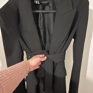 Svart kavaj från Zara - Säljer en stilren svart kavaj från Zara i storlek M. Kavajen har en klassisk design med knappar framtill och ett bälte för att ge en snygg passform. Perfekt för både vardag och fest! Använd endast ett fåtal gånger och är i nyskick. Pris går att diskuteras vid snabb affär. 