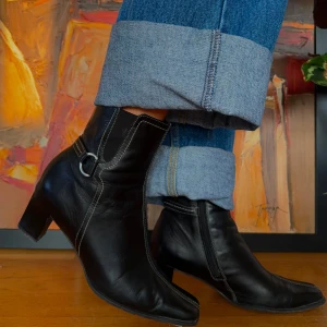 Svarta skinnstövletter med klack - Snygga svarta stövletter i skinn med en stilren design. De har en spetsig tå och en diskret klack som ger lite extra höjd. Stövletterna har dekorativa sömmar och en liten ringdetalj på sidan. Perfekta för både vardag och fest!