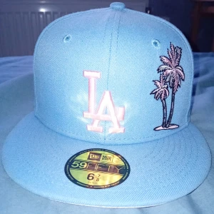 Ljusblå keps från New Era - Säljer en supercool ljusblå keps från New Era. Den har en broderad LA-logga och palmer på framsidan, vilket ger den en skön sommarvibe. På baksidan finns en liten taco-brodering som ger en rolig twist. Kepsen är i 59FIFTY-modell och passar perfekt för alla som älskar streetwear.