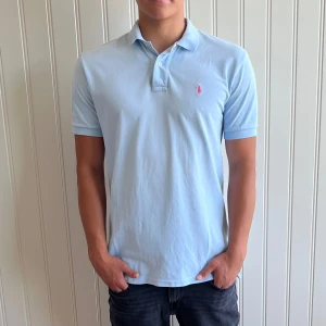 Ljusblå pikétröja från Ralph Lauren - Säljer en snygg ljusblå pikétröja från Ralph Lauren. Den har en klassisk krage med knappar och det ikoniska rosa broderade logotypen på bröstet. Perfekt för en stilren och avslappnad look. Passar bra till både vardag och fest!