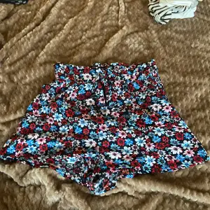 Säljer ett par supersöta blommiga shorts från H&M Divided. De är i storlek 34 och har ett färgglatt mönster med blommor i rött, blått och rosa på en svart bakgrund. Shortsen har en hög midja och en bekväm passform, perfekta för sommardagar! 🌸