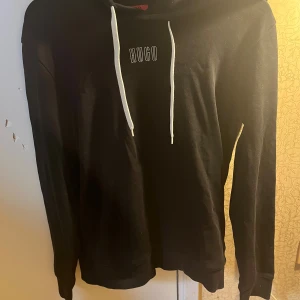 Svart hoodie från Hugo - Säljer en snygg svart hoodie från Hugo Boss. Den har en cool vit logga både fram och bak, och vita snören i huvan. Perfekt för en casual look eller när du bara vill chilla. Unisex-modell som passar alla!