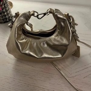 Guldig lite väska  - Väska som går att ha både i handen, armvecket samt kommer den med ett matchade långt band till.  Använd men i fint skick.  Köpt för 900kr