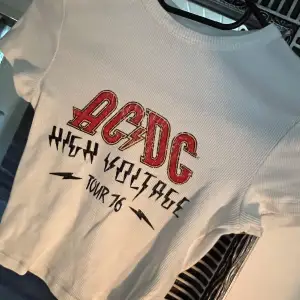 Säljer en supercool vit AC/DC T-shirt från Bershka. Den har ett tryck med 'High Voltage Tour 76' i rött och svart på framsidan. T-shirten är ribbad och kortärmad, perfekt för en rockig look! 🎸