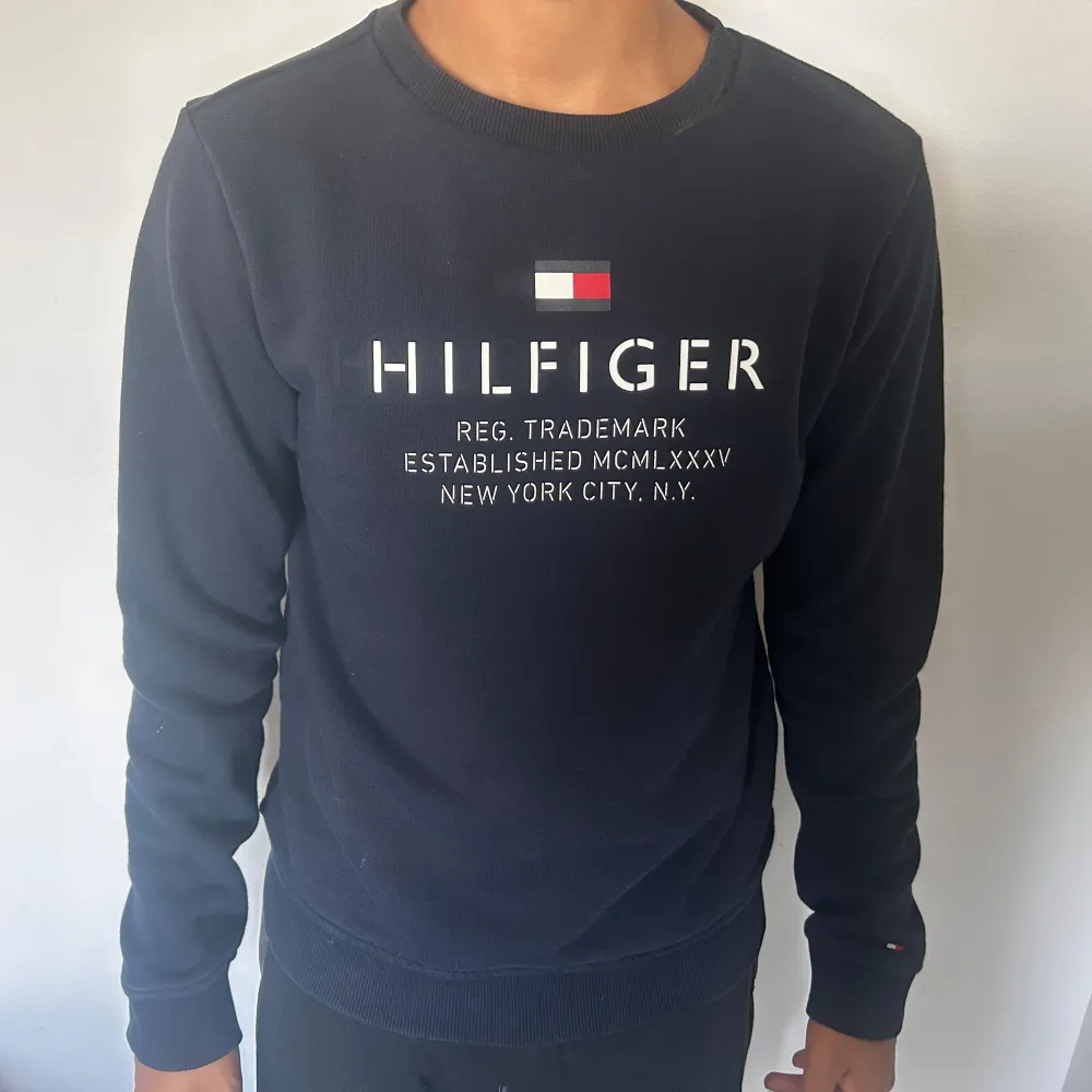 Säljer en snygg mörkblå tröja från Tommy Hilfiger. Den har ett klassiskt tryck på framsidan med texten 'HILFIGER' och detaljer i rött, vitt och blått. Tröjan är långärmad och har ribbade muddar vid ärmslut och nederkant. Perfekt för en casual look!. Tröjor & Koftor.