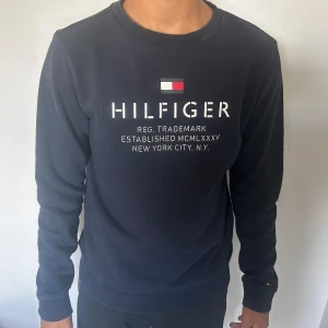 Mörkblå tröja från Tommy Hilfiger - Säljer en snygg mörkblå tröja från Tommy Hilfiger. Den har ett klassiskt tryck på framsidan med texten 'HILFIGER' och detaljer i rött, vitt och blått. Tröjan är långärmad och har ribbade muddar vid ärmslut och nederkant. Perfekt för en casual look!