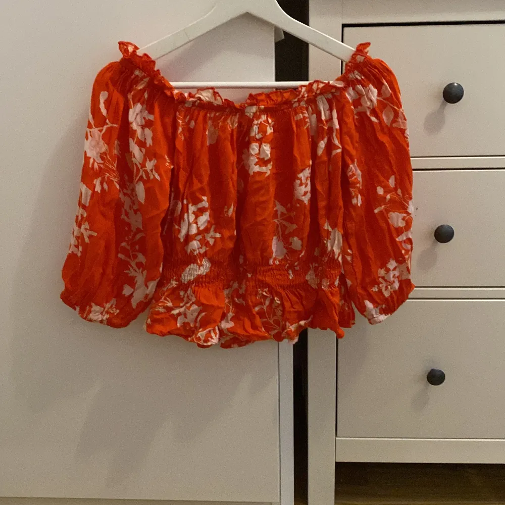 Säljer en superfin orange offshoulder topp från BikBok med vita blommor. Den har en elastisk kant upptill och vid midjan för en bekväm passform. Perfekt för sommardagar eller en festlig kväll!. Toppar.