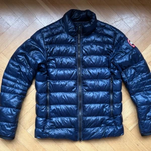 Blå Crofton dunjacka från Canada Goose - L - Säljer en snygg blå dunjacka från Canada Goose med broderad logga på ärmen, i storlek L. Jackan är perfekt för kyliga dagar. Den är långärmad och har en klassisk pufferdesign. Säljs i butik för 9950 SEK. Utmärkt skick, med två saker att notera: 1) litet hål i tyget, täckt av en särskild tyglapp. Se sista bilden. Märks inte när man bär den. 2) Hängslen försvann under vår flytt (har aldrig använt dem själv 🤷‍♂️)