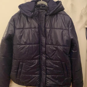 Mörkblå pufferjacka med avtagbar huva - Säljer en snygg mörkblå pufferjacka med avtagbar huva. Jackan har en quiltad design och är perfekt för kyligare väder. Den har långa ärmar och stängs med dragkedja samt tryckknappar.  Perfekt för vintern!