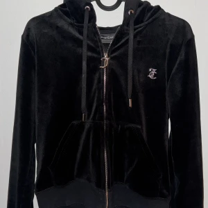 Svart Set från Juicy Couture - Snygg svart velour hoodie från Juicy Couture med glittrande stenar som bildar texten 'Juicy' på ryggen. Tröjan har dragkedja och huva med snören. Svarta matchande byxor med ”Juicy” på rumpan. Perfekt för en avslappnad och stilren look.